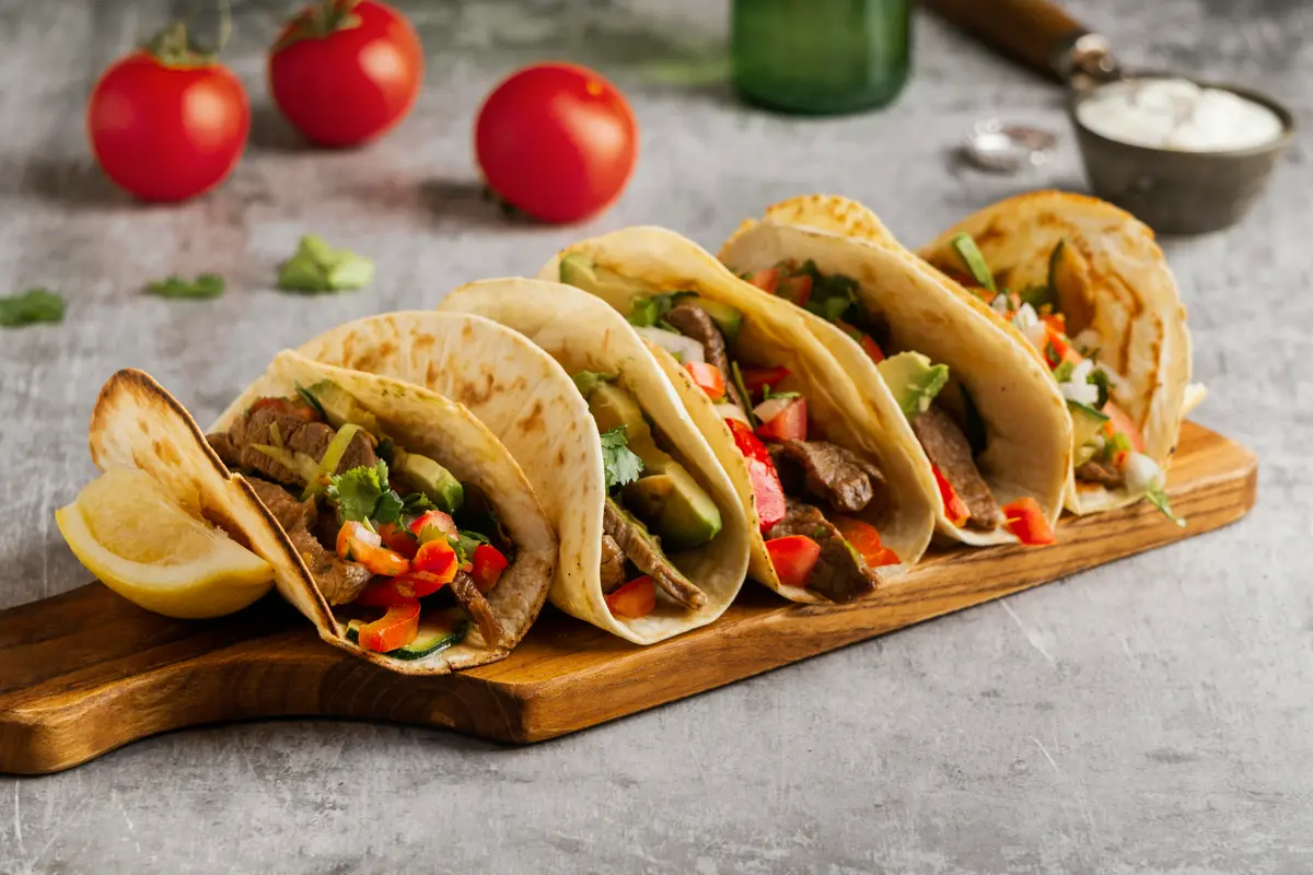 Tacos de lengua served on a wooden plate with fresh cilantro, diced onions, and lime wedges.